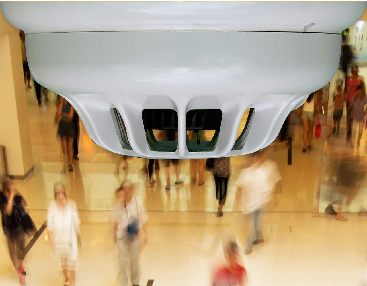 A smoke detector installed in a commercial building, with people walking below