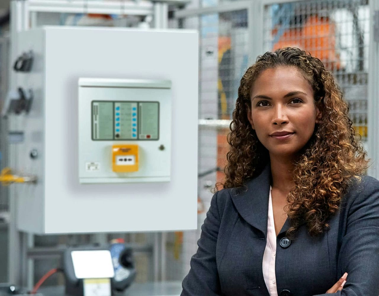 A woman dressed in business formals standing with her arms folded, near a fire control panel